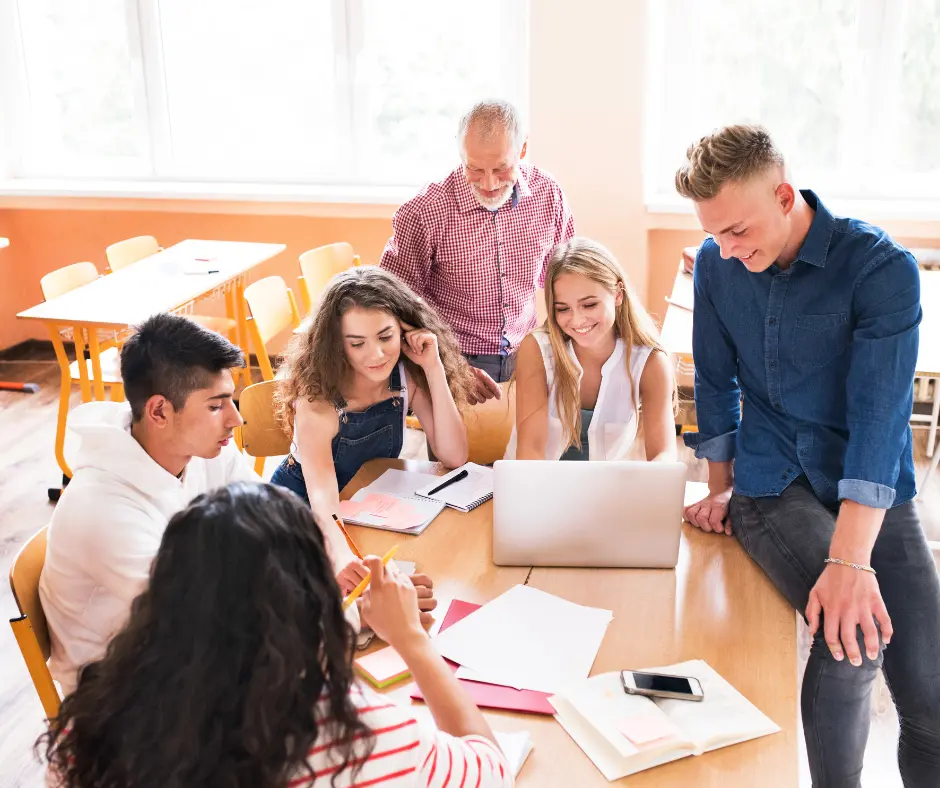 Mehrere Personen sitzen und stehen um einen Tisch mit Laptop und Schreibmaterialien und konzipieren Ideen