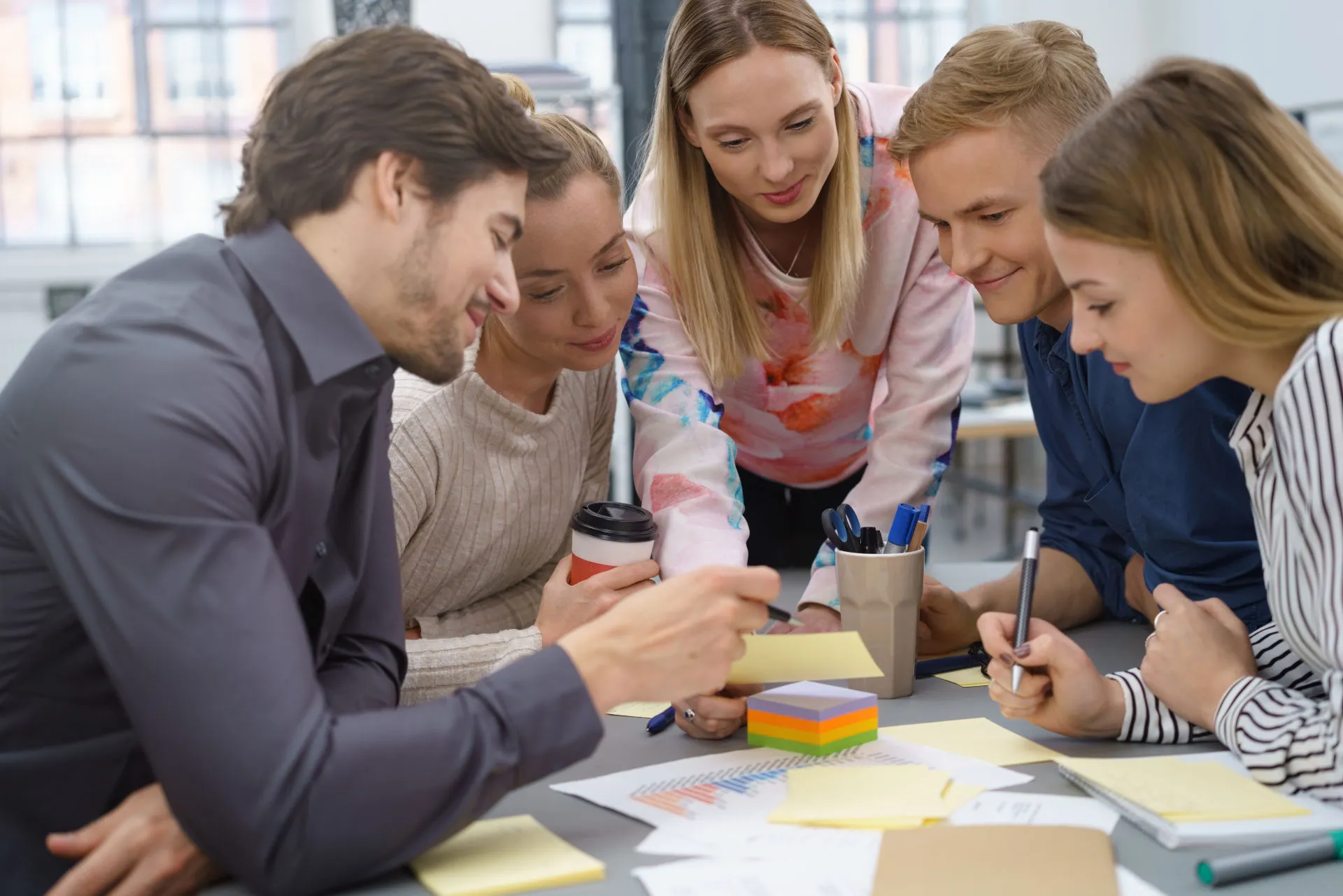 Fünf Personen mit Stiften und Kaffee in der Hand sitzen dicht beieinander am Tisch über Klebezetteln und konzipieren Ideen